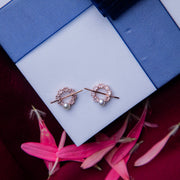 A pair of rose gold-colored stud earrings displayed on a blue cushion with a floral pattern, surrounded by pink petals.